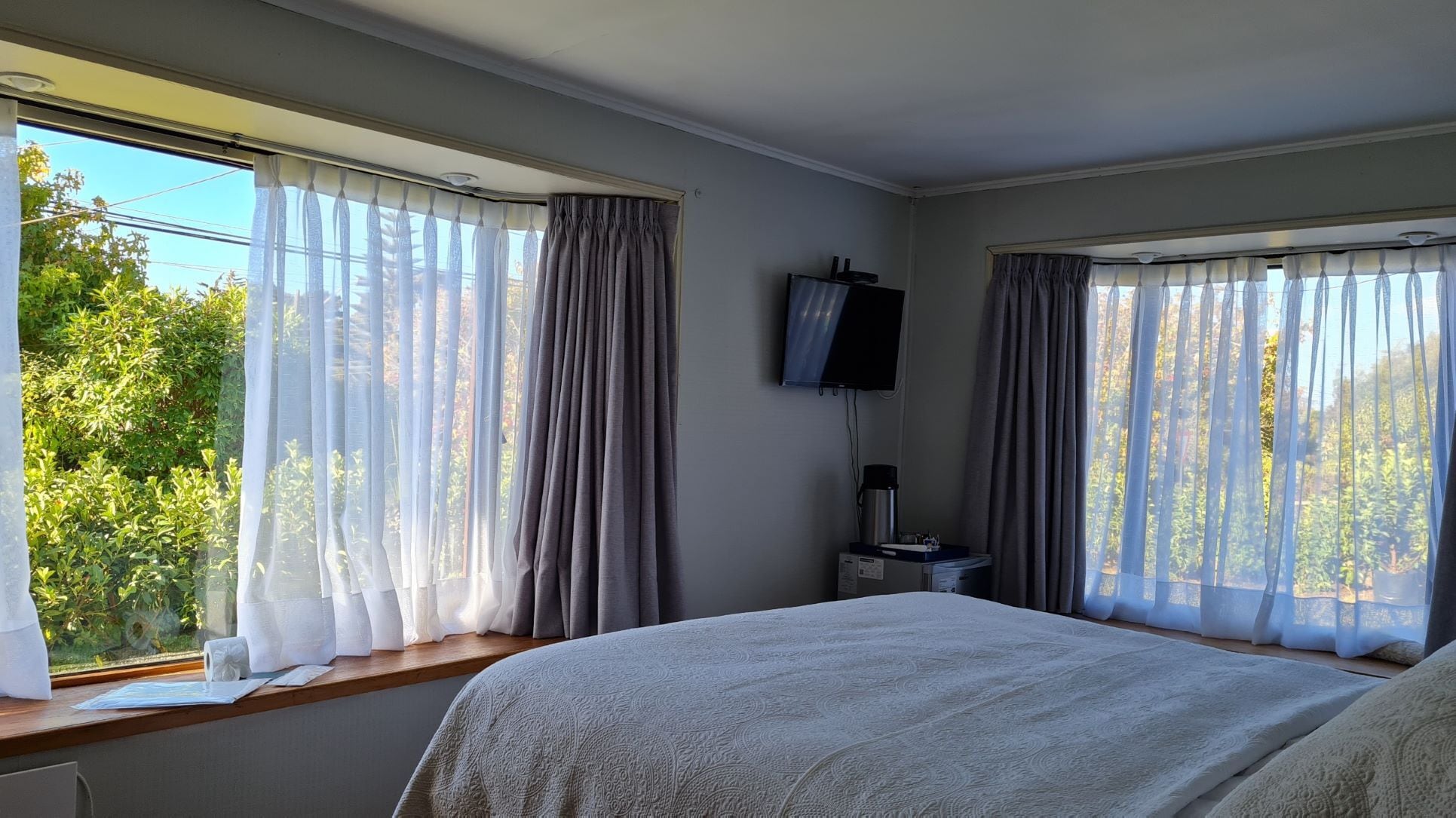 Un dormitorio con una cama grande, dos ventanas con cortinas traslúcidas, un televisor de pantalla plana y una mesita de noche pequeña.
