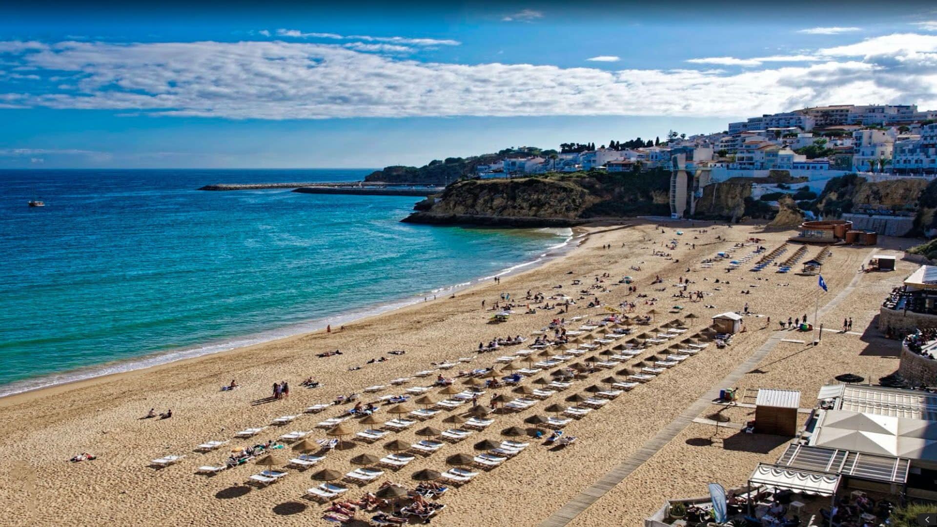 Une plage bordée de rangées de transats et de parasols, un littoral rocheux et des bâtiments en arrière-plan.