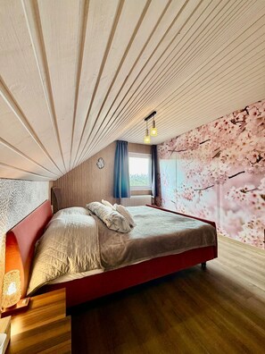 Une chambre avec un plafond en pente, un lit avec une tête de lit rouge et un mur recouvert d’un papier peint à motifs de fleurs de cerisier.