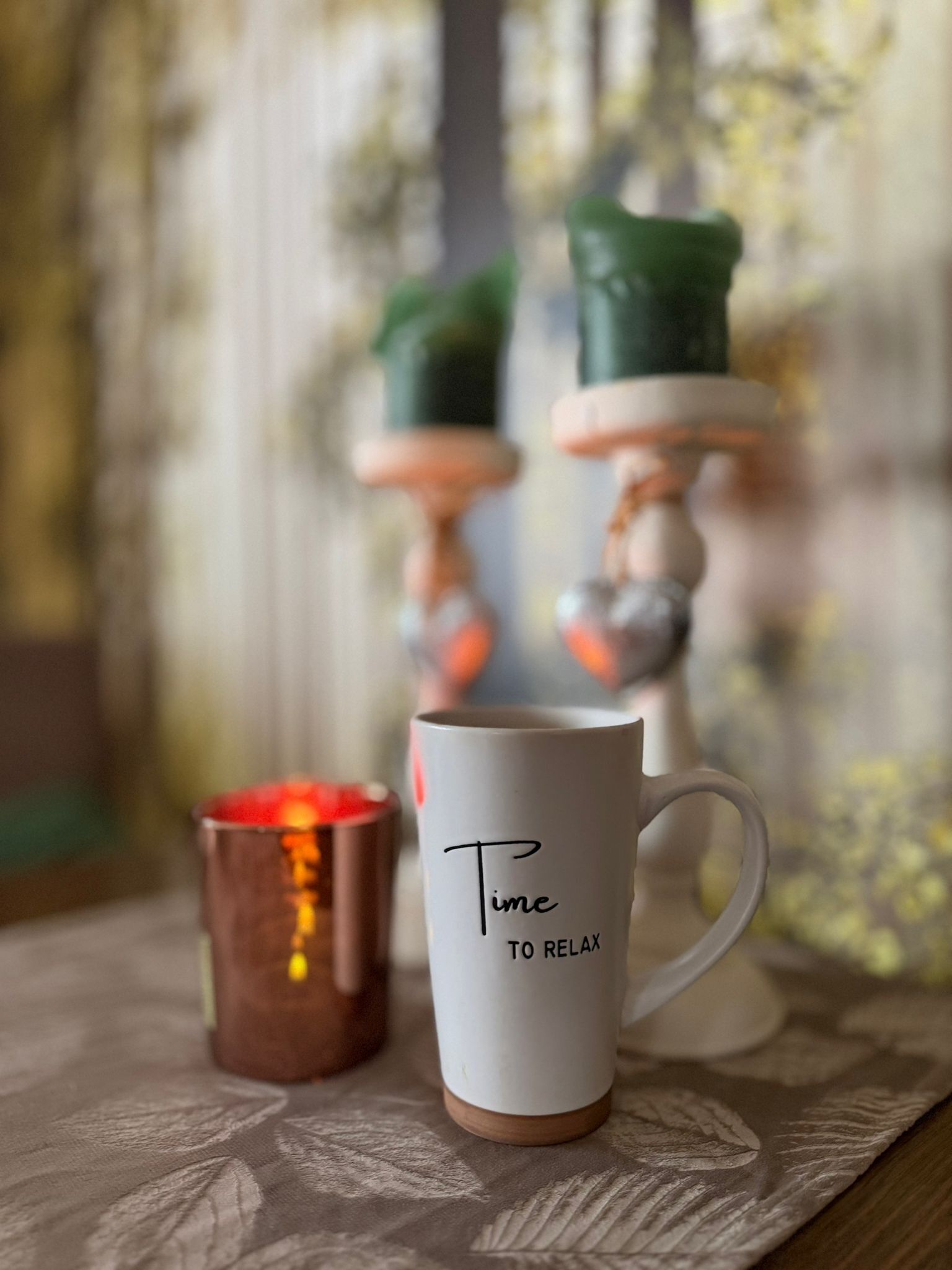 A white mug with 'Time to Relax' text, a copper candle holder, and a decorative candle on a table.