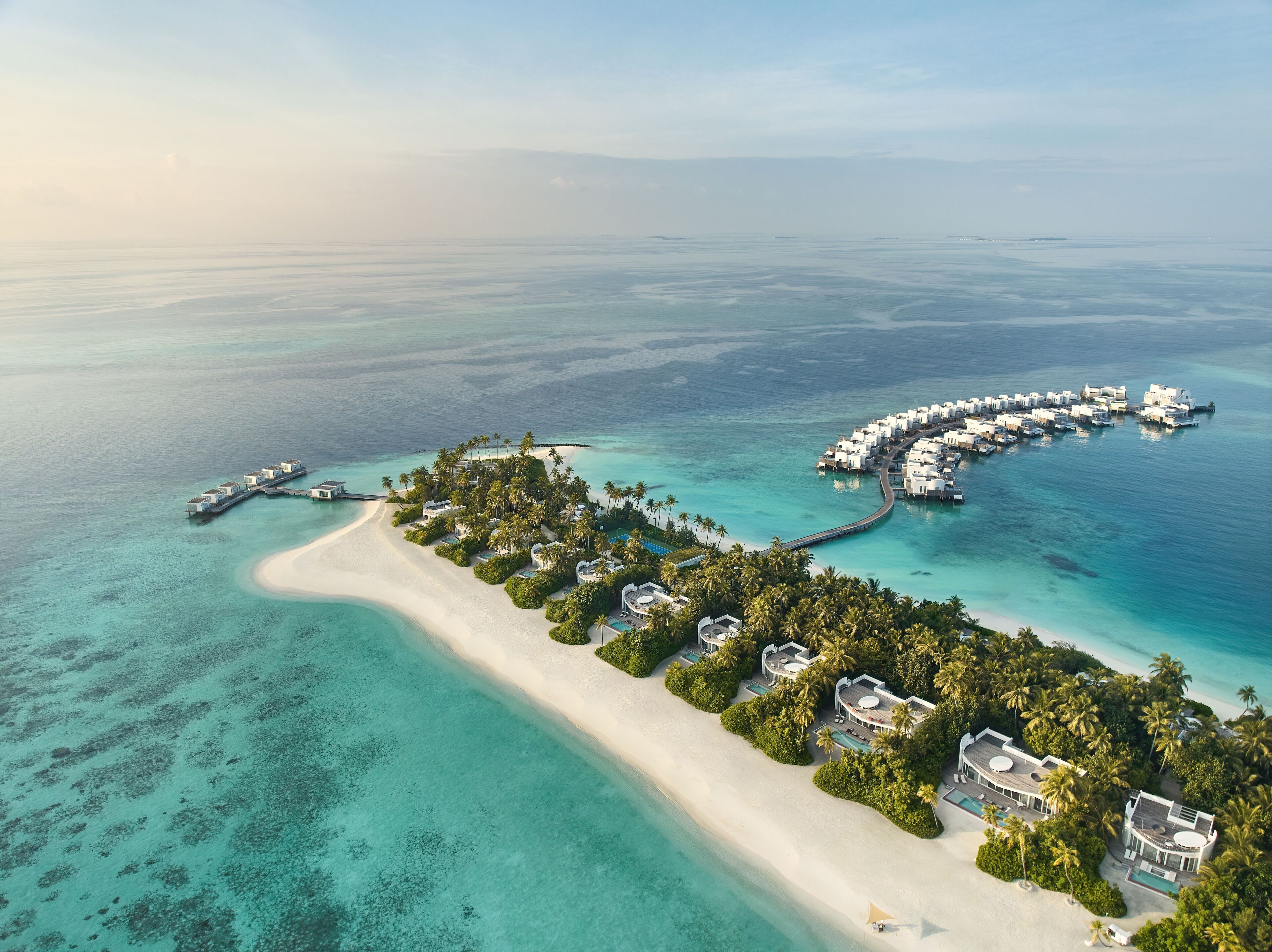 Vista aérea de un resort con bungalows sobre el agua, una playa de arena y aguas turquesas cristalinas.