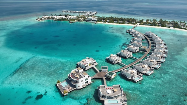 An aerial view of a resort with overwater bungalows connected by walkways, clear turquoise waters, and a distant island with a pier. - Jumeirah Olhahali Island  (Olhahali)