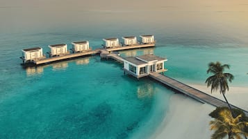 A series of overwater bungalows connected by a wooden walkway, with clear turquoise water and a palm tree visible.