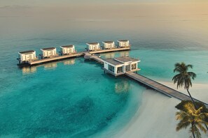 Une série de bungalows sur pilotis reliés par une passerelle en bois, avec une eau turquoise cristalline et un palmier visible.