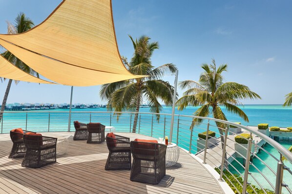 A wooden deck with wicker chairs and a table, overlooking a clear blue sea and palm trees.