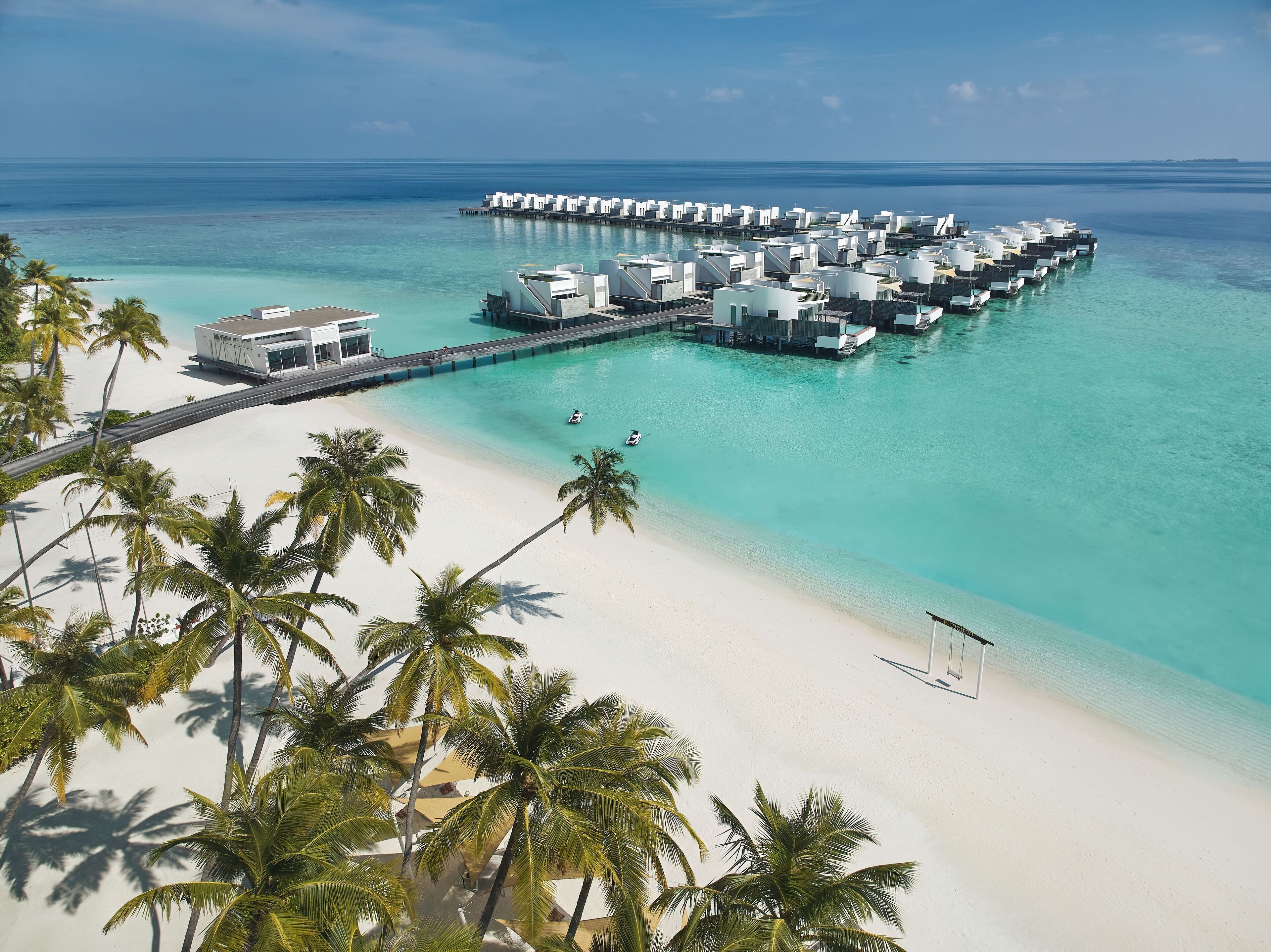 Un resort sobre el agua con varias cabañas conectadas por pasarelas a una playa de arena y palmeras.