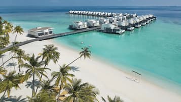 Un resort sobre el agua con varias cabañas conectadas por pasarelas a una playa de arena y palmeras.