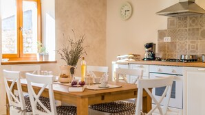 Un coin repas avec une table en bois, des chaises blanches ornées de coussins à motifs, un vase contenant des plantes séchées et, au fond, une cuisine.