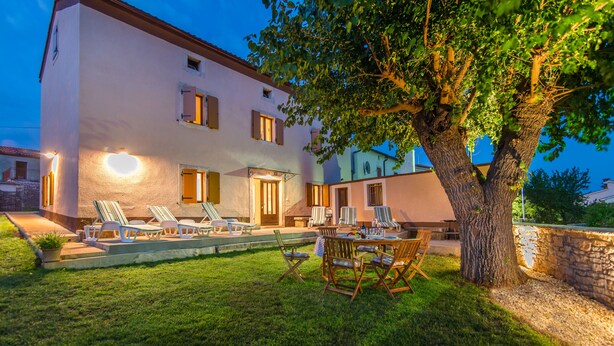 Un bâtiment à deux étages doté d’une pelouse bien entretenue, d’un espace extérieur aménagé avec des chaises et des tables, et d’un grand arbre qui offre de l’ombre.