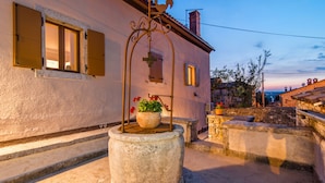 Un puits en pierre, avec un pot de fleurs devant un bâtiment orné d’une croix sur le mur et doté d’une fenêtre avec des volets.