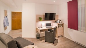 Une chambre moderne avec un bureau en bois, un ordinateur portable, une télévision, un canapé gris et une fenêtre avec un store rouge.