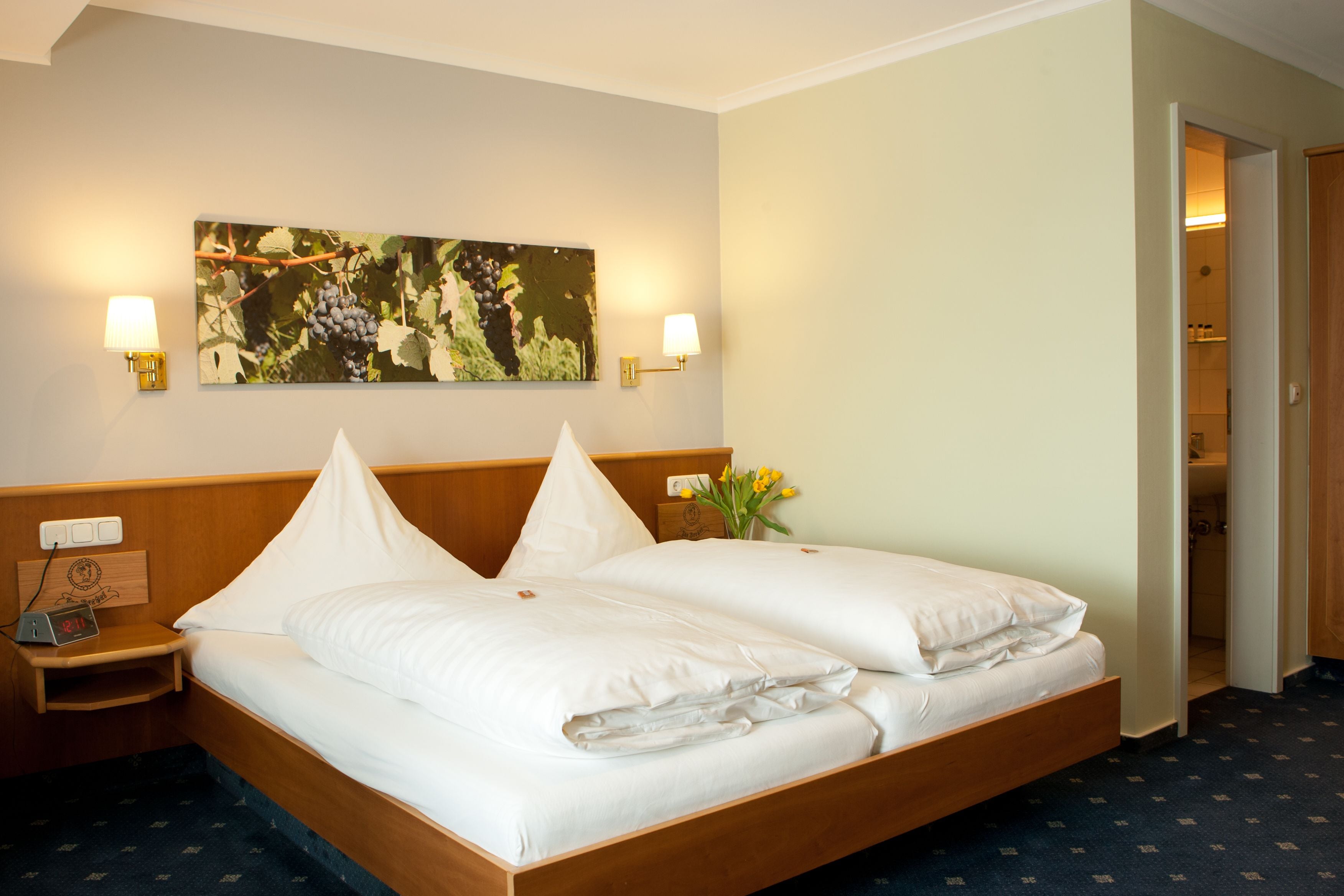 A double bed with white linens, two bedside tables with lamps, a wall-mounted painting of grapes, and a wooden headboard.