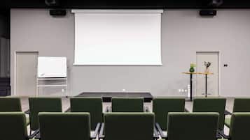 A conference room with a projector screen, green chairs, a whiteboard, and a small table with a vase of flowers.
