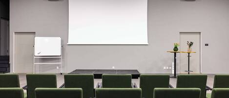A conference room with a projector screen, green chairs, a whiteboard, and a small table with a vase of flowers.