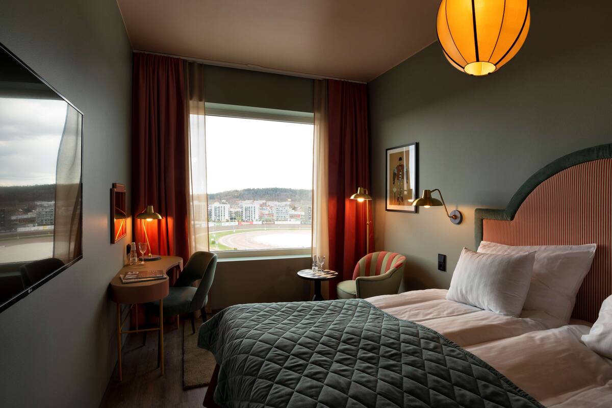 A hotel room with a large bed, a desk with a chair, a small table, and a view of the cityscape through the window.