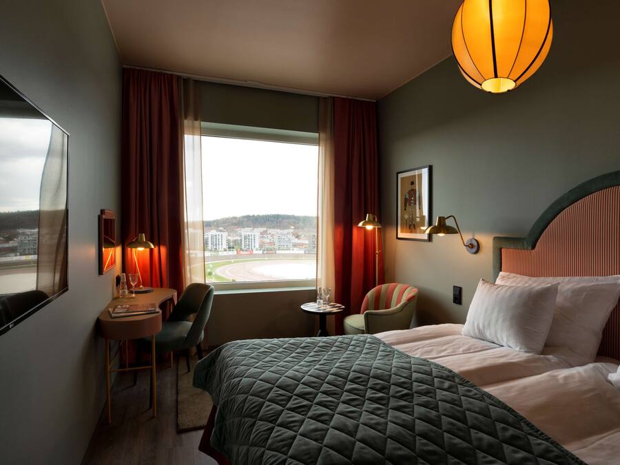 Habitación de hotel con una cama grande, un escritorio con silla, una mesita y vistas a la ciudad desde la ventana.