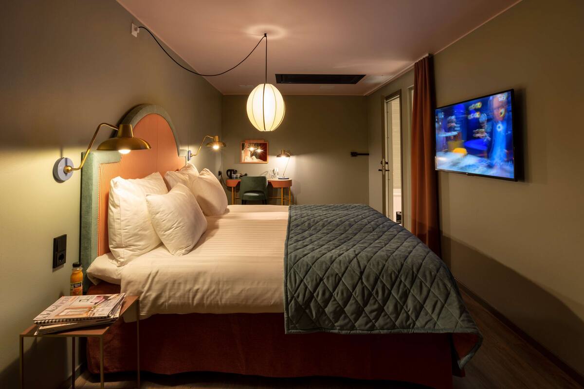 A hotel room with a bed, bedside lamps, a desk, a chair, a television, and a door.