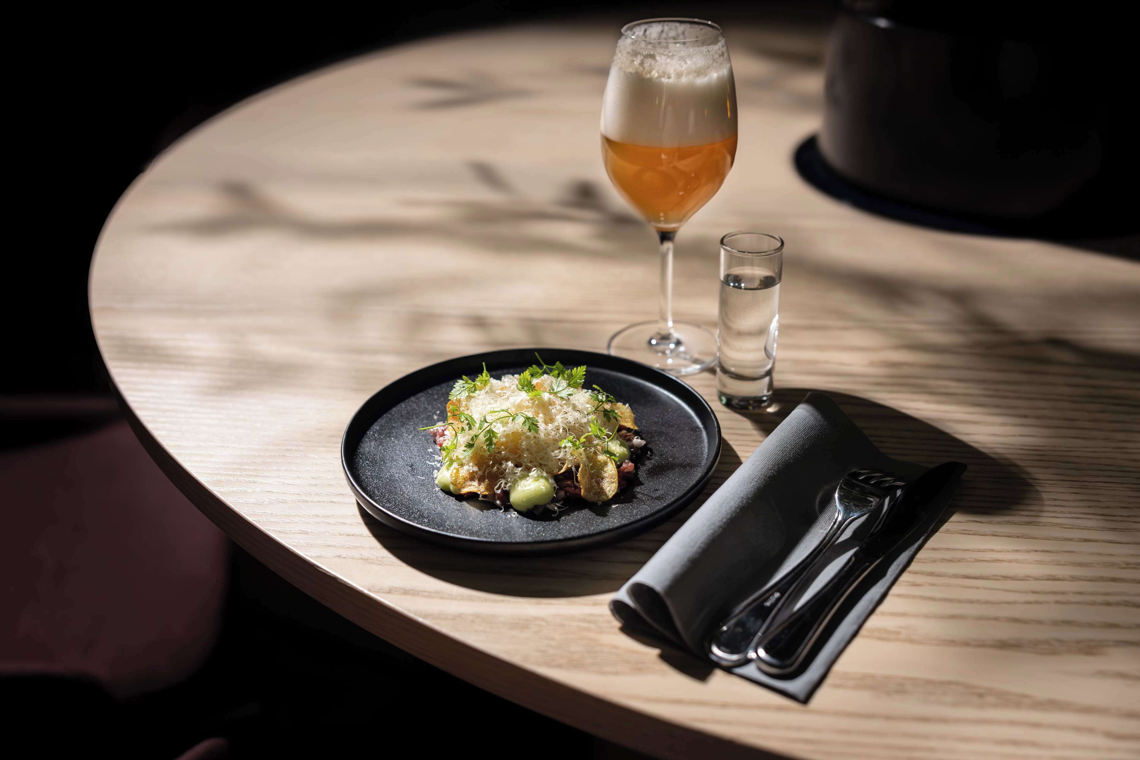 Una mesa redonda de madera con un plato de comida negro, una jarra de cerveza y un vaso de agua.