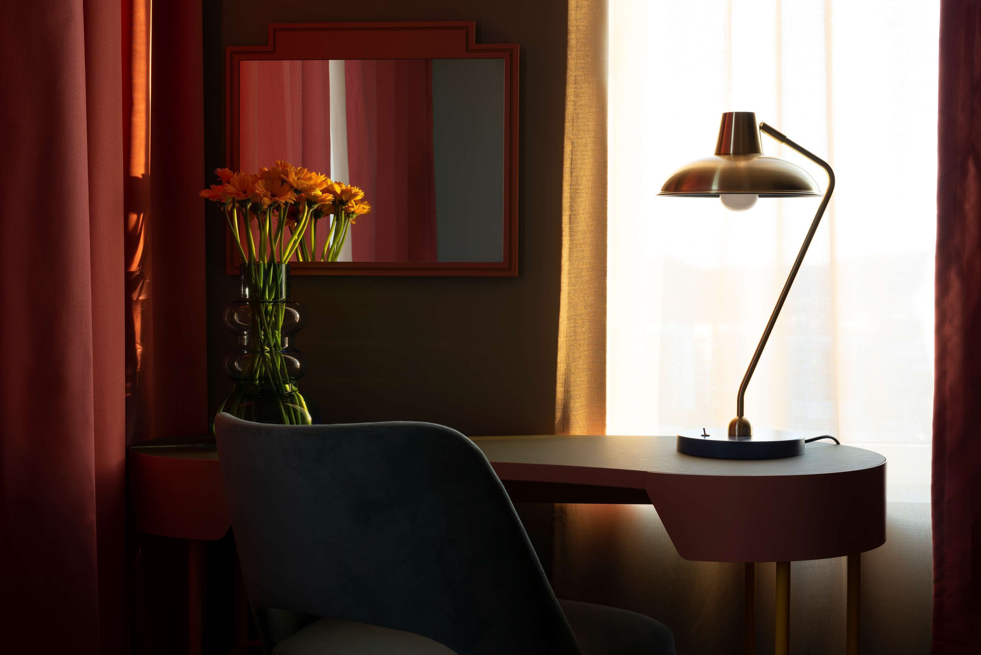 A room with a desk, chair, lamp, and a vase with flowers.
