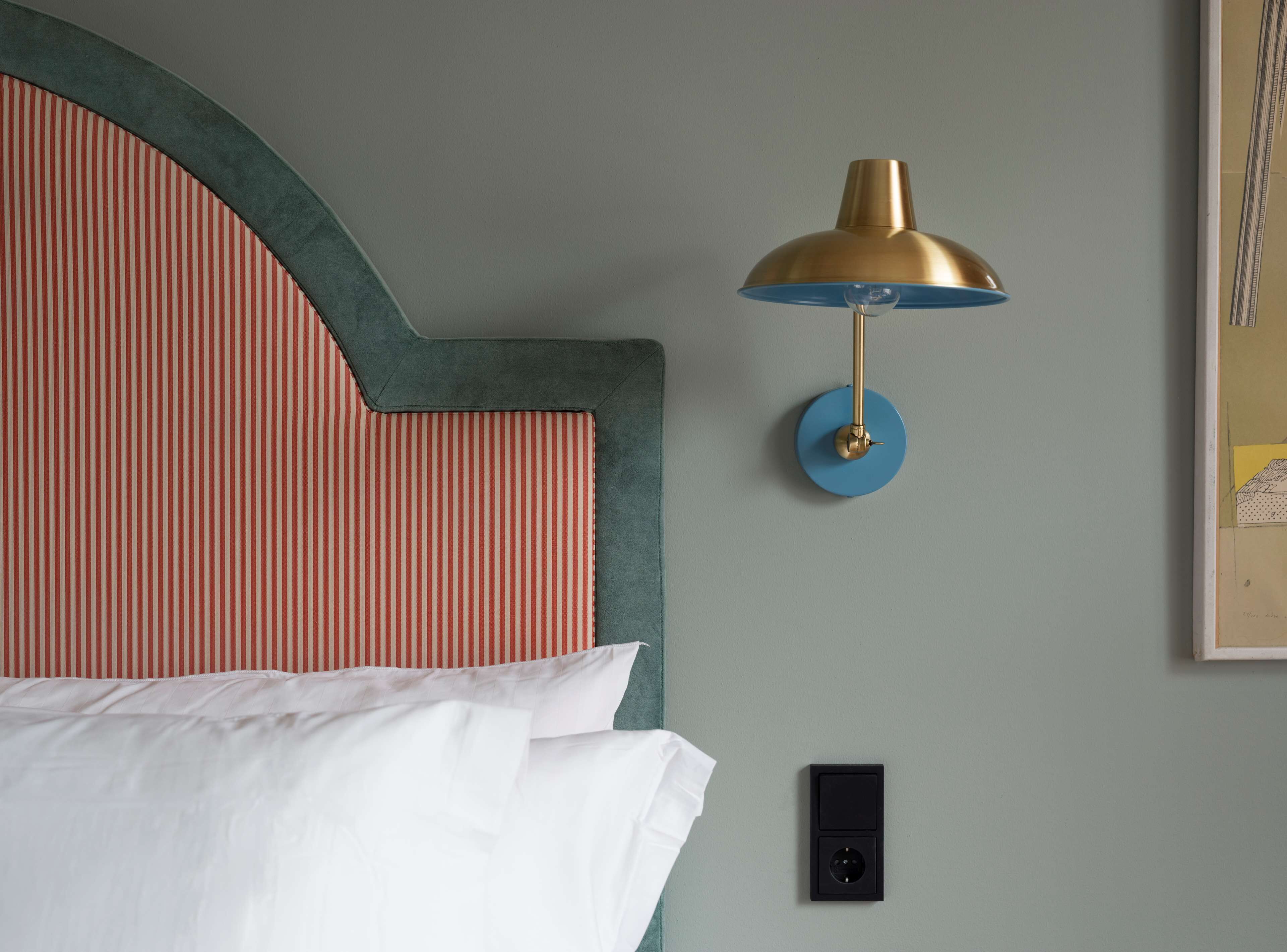 Una cama con ropa blanca, un cabecero a rayas, una lámpara azul fijada a la pared y un enchufe eléctrico negro.
