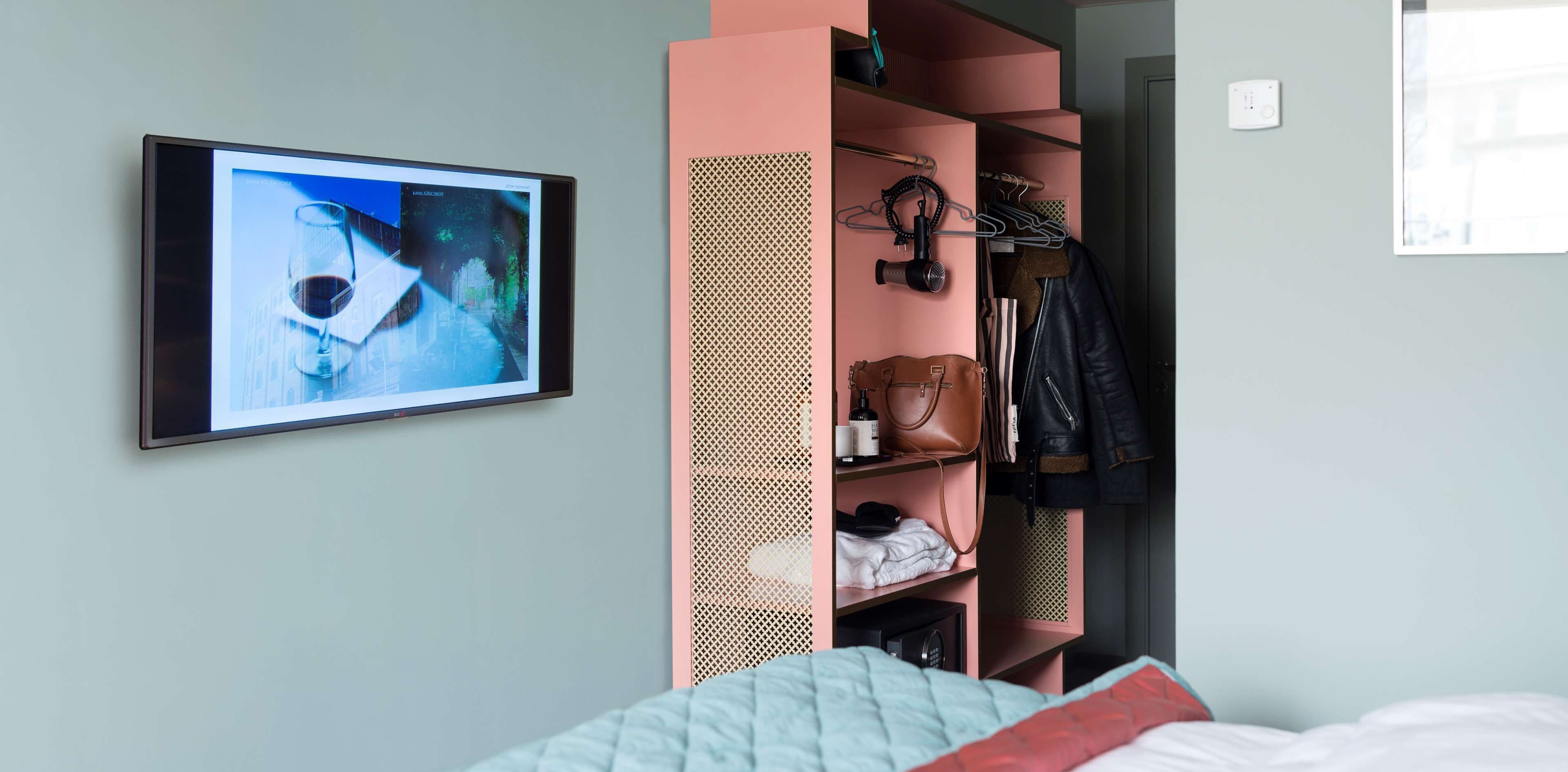 A bedroom with a bed, a TV mounted on the wall, a pink wardrobe, and a coat rack.