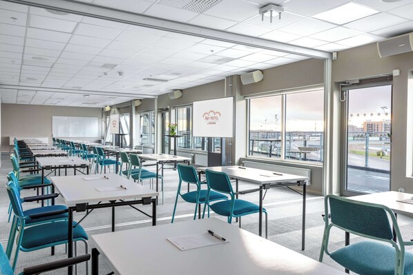 Une salle de conférence avec des rangées de tables et de chaises, de grandes fenêtres donnant sur la ville et un tableau blanc au mur.
