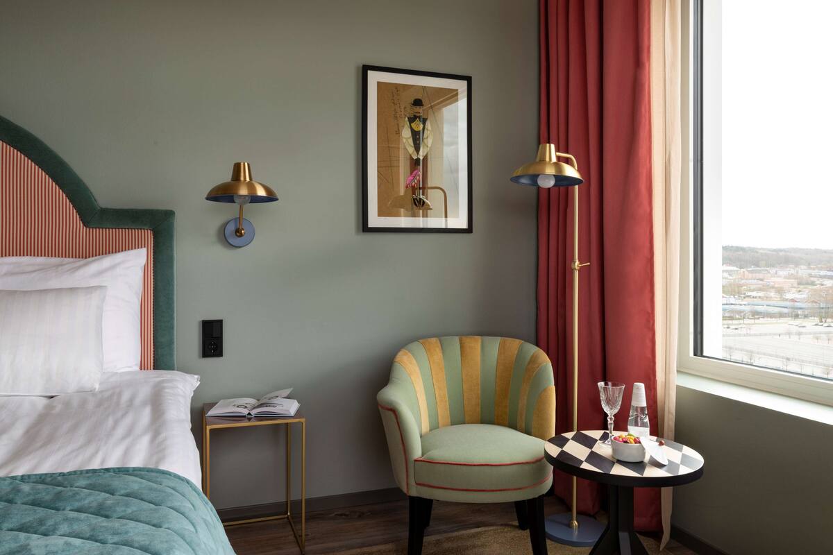 A hotel room with a bed, a chair, a small table with a glass, and a window with curtains.