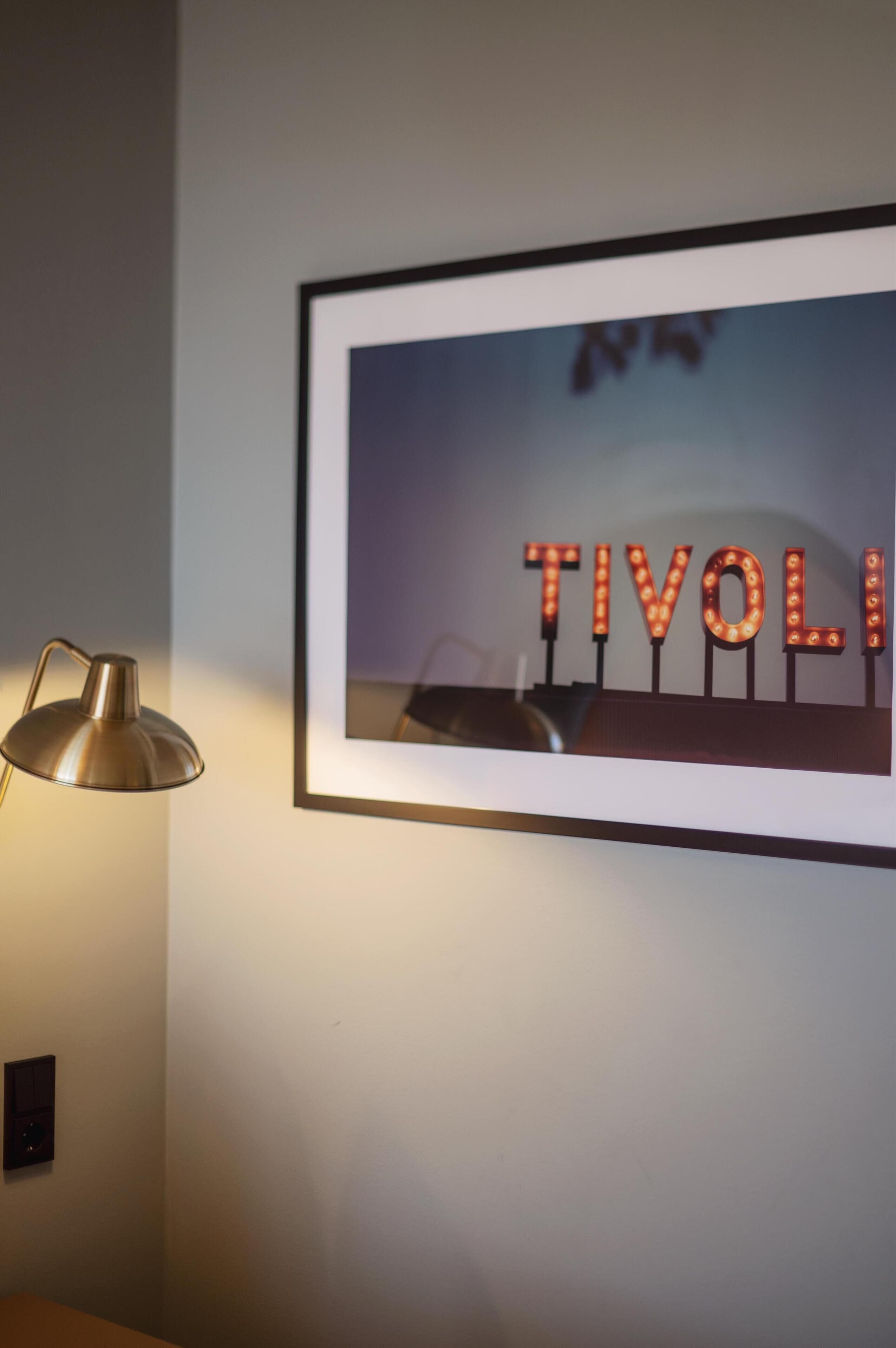 Un cuadro enmarcado con letras iluminadas que deletrean "TIVOLI" en una pared, una lámpara a la izquierda y un tablero de interruptores debajo.