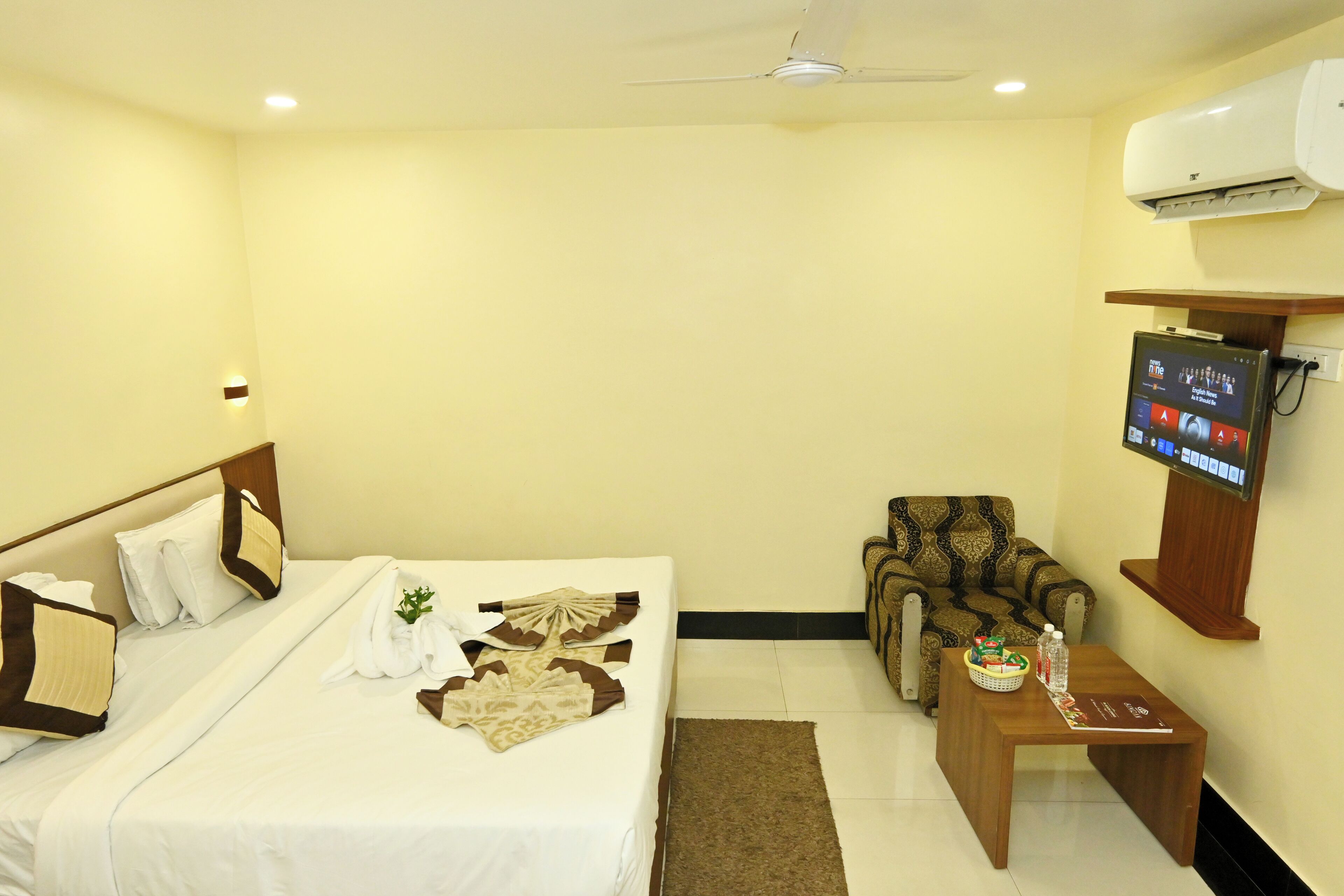 Habitación de hotel con cama, una silla, una mesita con control remoto y un televisor montado en la pared.