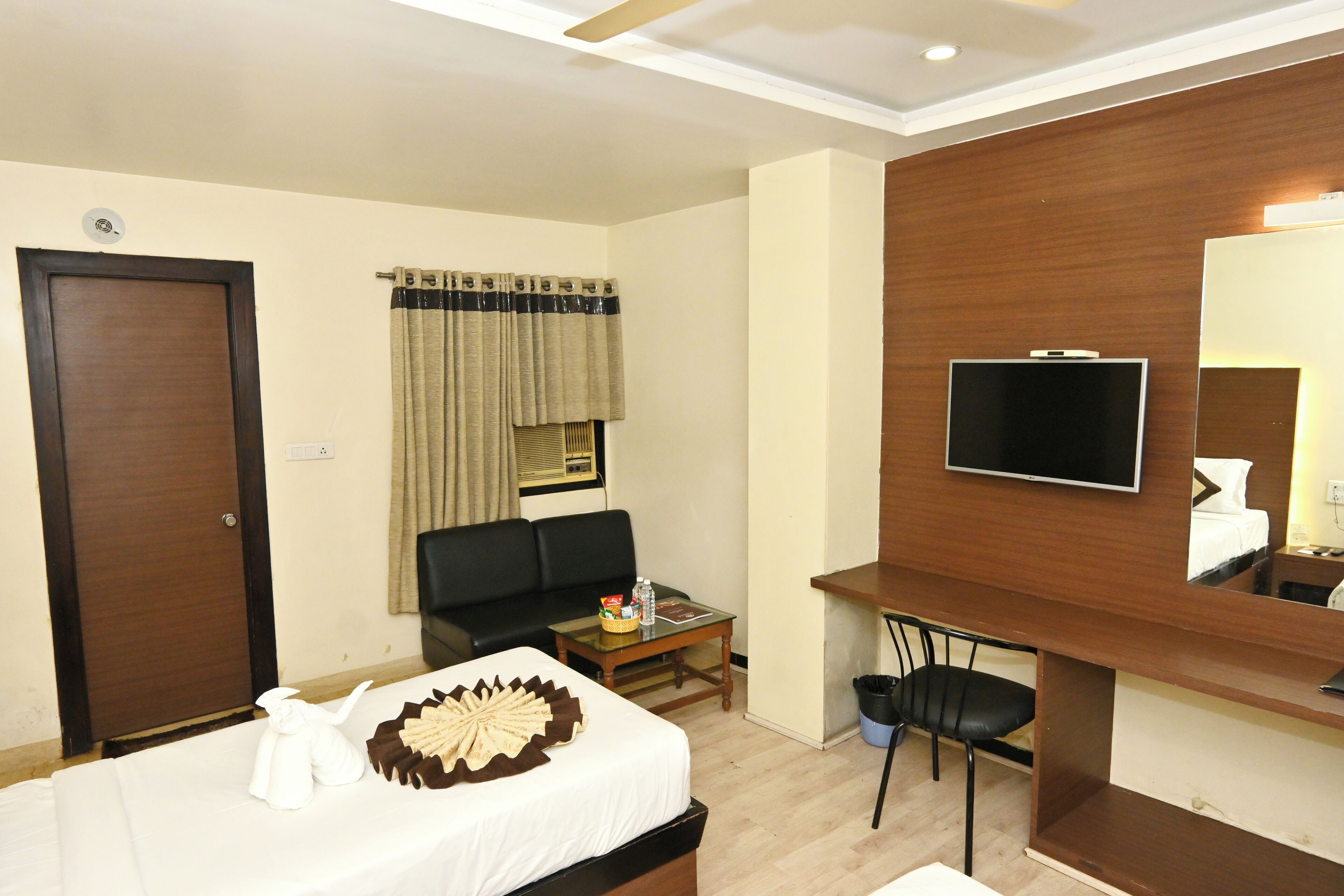 A hotel room with a bed, a TV, a desk, a chair, a sofa, and a window with curtains.