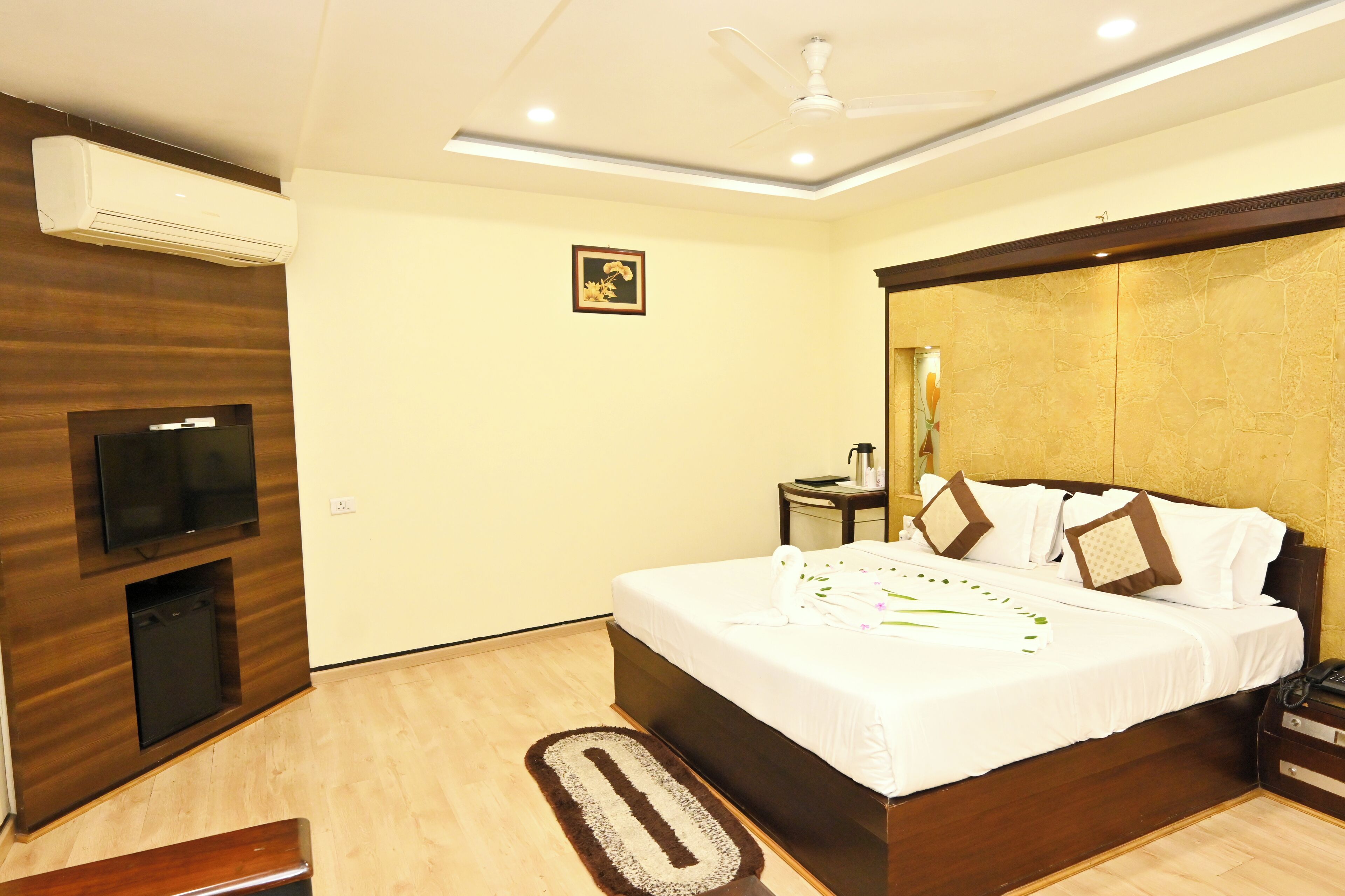 A modern hotel room with a wooden accent wall, a flat-screen TV, a bed with white bedding and decorative pillows, a bedside table with a lamp, and a ceiling fan.