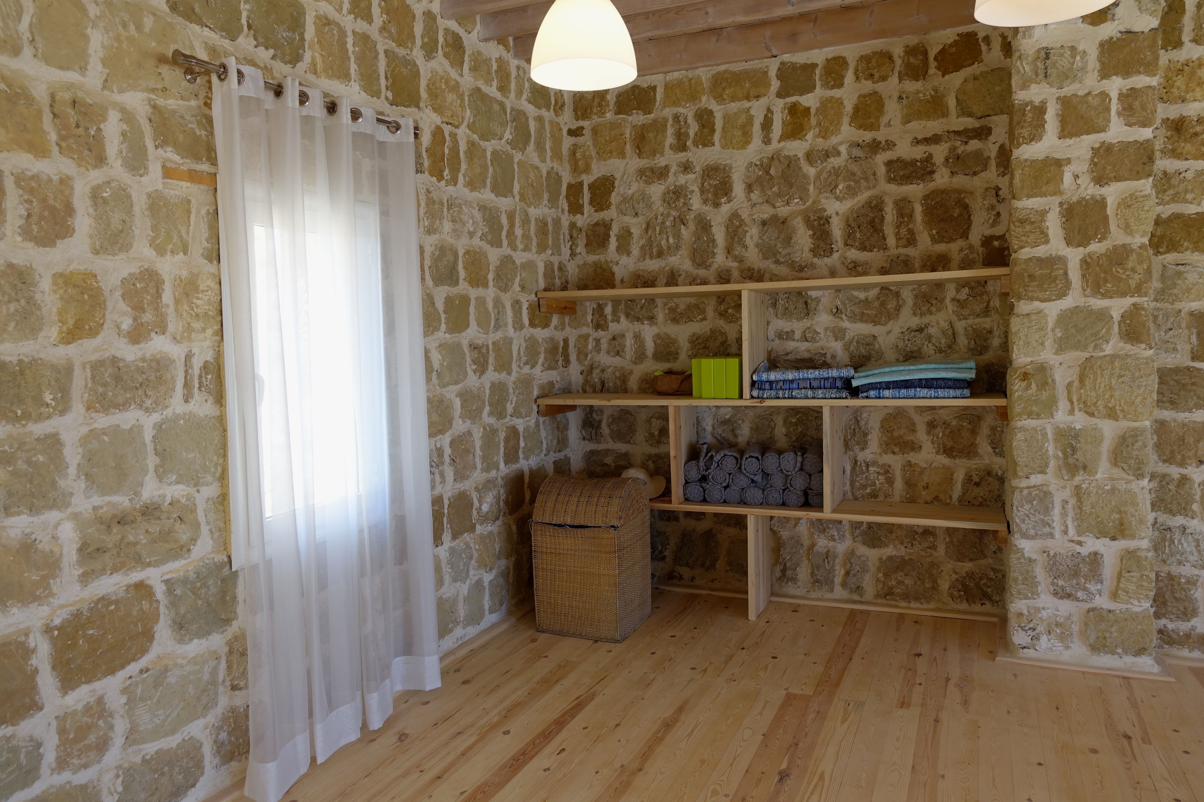 Una habitación con paredes de piedra, suelo de madera, una cortina blanca y estantes con cestas y mantas dobladas.