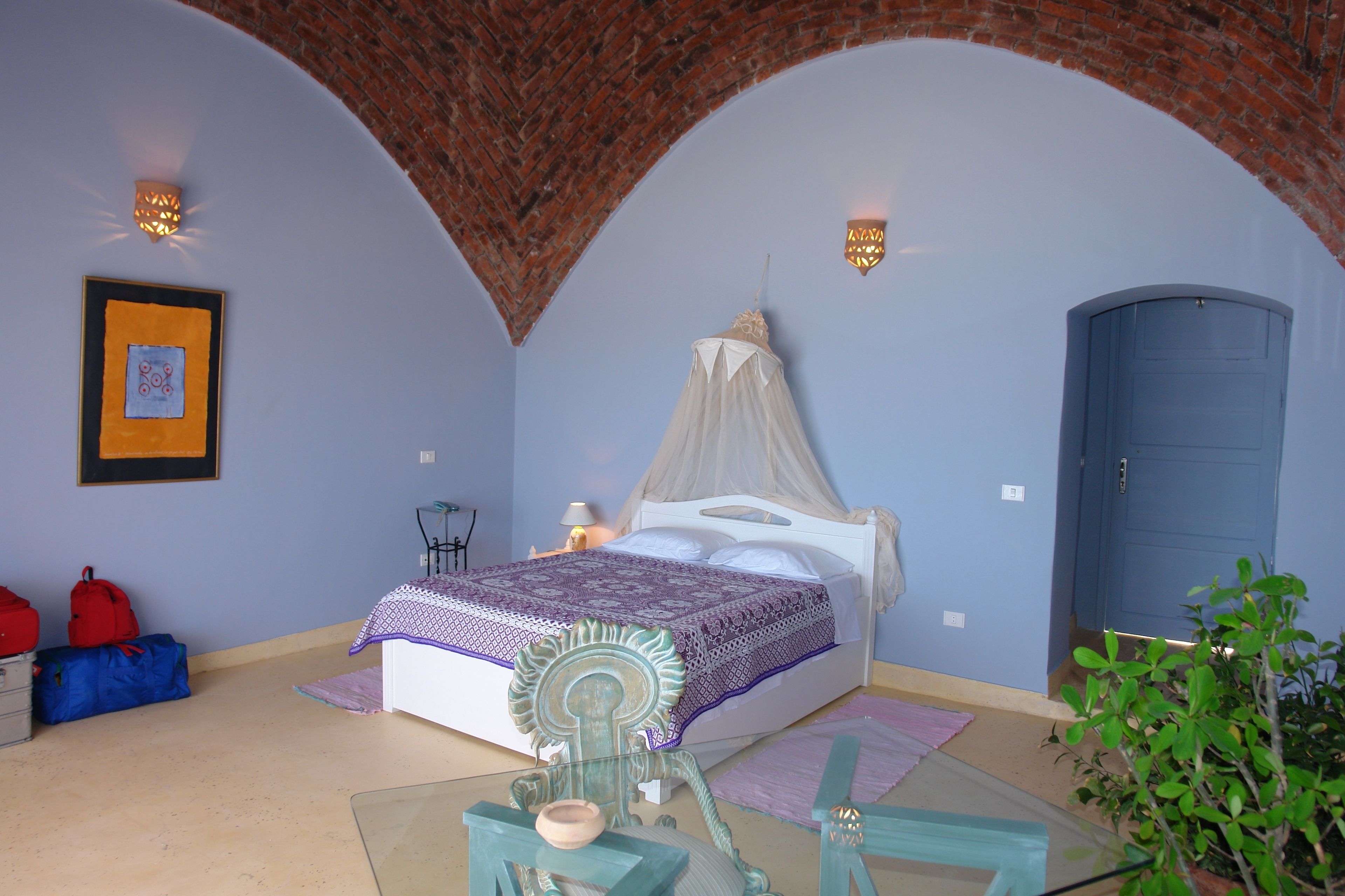 Un dormitorio con una cama bajo un dosel blanco, una puerta azul y un arco de ladrillo.