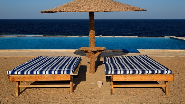Zona de piscina frente a la playa con dos sillones de madera y un toldo de paja.