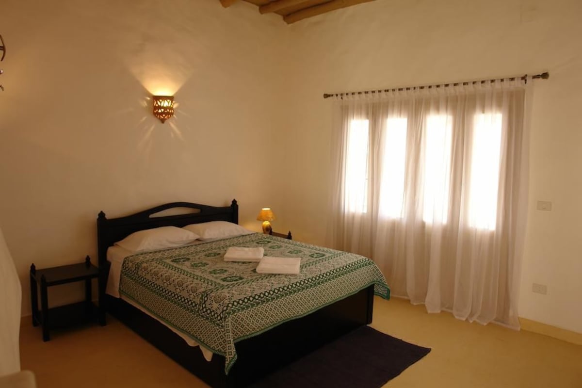 Un dormitorio con cama, mesita de noche, lámpara de pared y ventana con cortinas.
