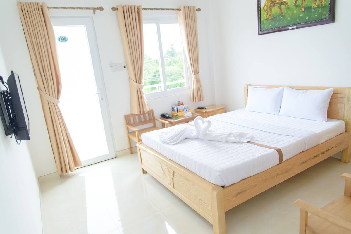 A bedroom with a wooden bed, white bedding, a desk with a chair, and a window with curtains.