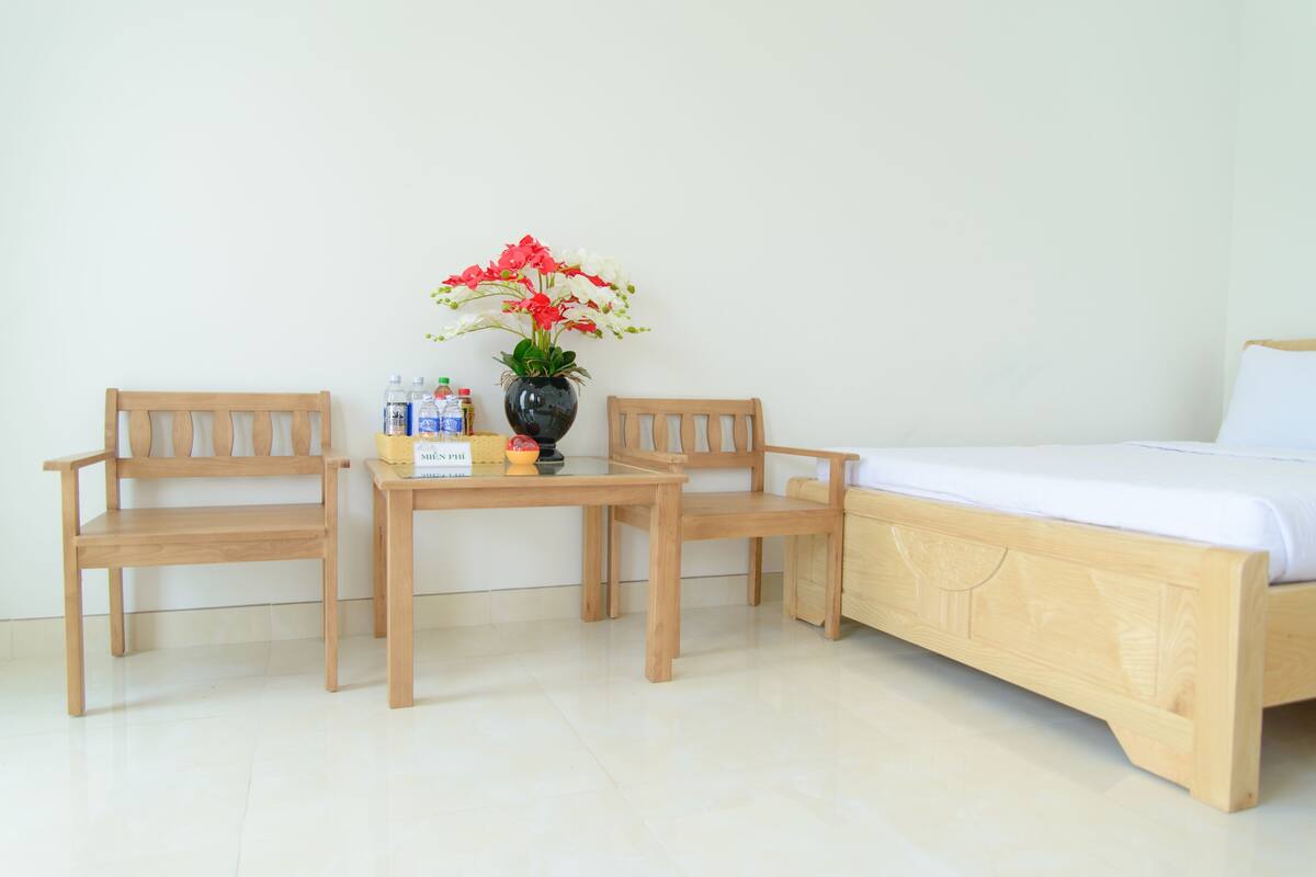 A room with a wooden bed, two wooden chairs, a small table, a vase with flowers, and a few bottles.