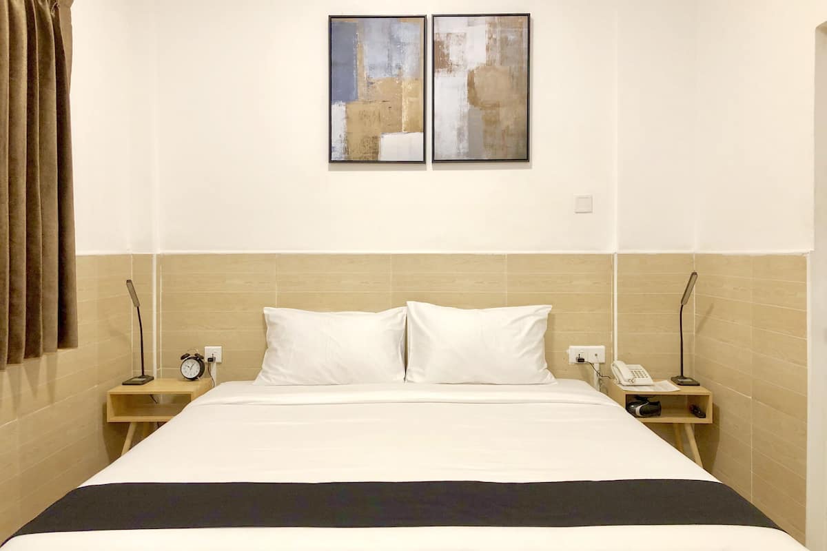 Quarto de hotel com uma cama grande, duas mesas de cabeceira, um criado-mudo com telefone e despertador, e duas pinturas abstratas na parede.