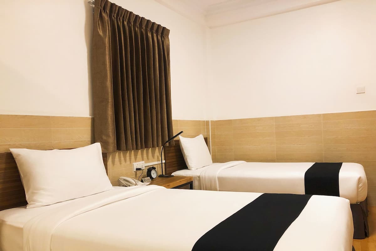 Quarto de hotel com duas camas, uma mesa de cabeceira com telefone e relógio, e uma janela com cortinas.