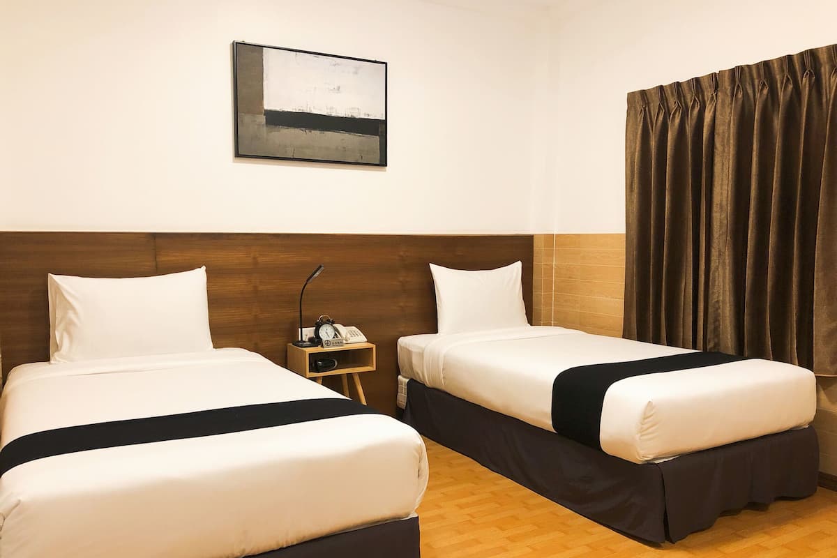 Quarto de hotel com duas camas, mesa de cabeceira com abajur e telefone, quadro na parede e janela com cortinas.