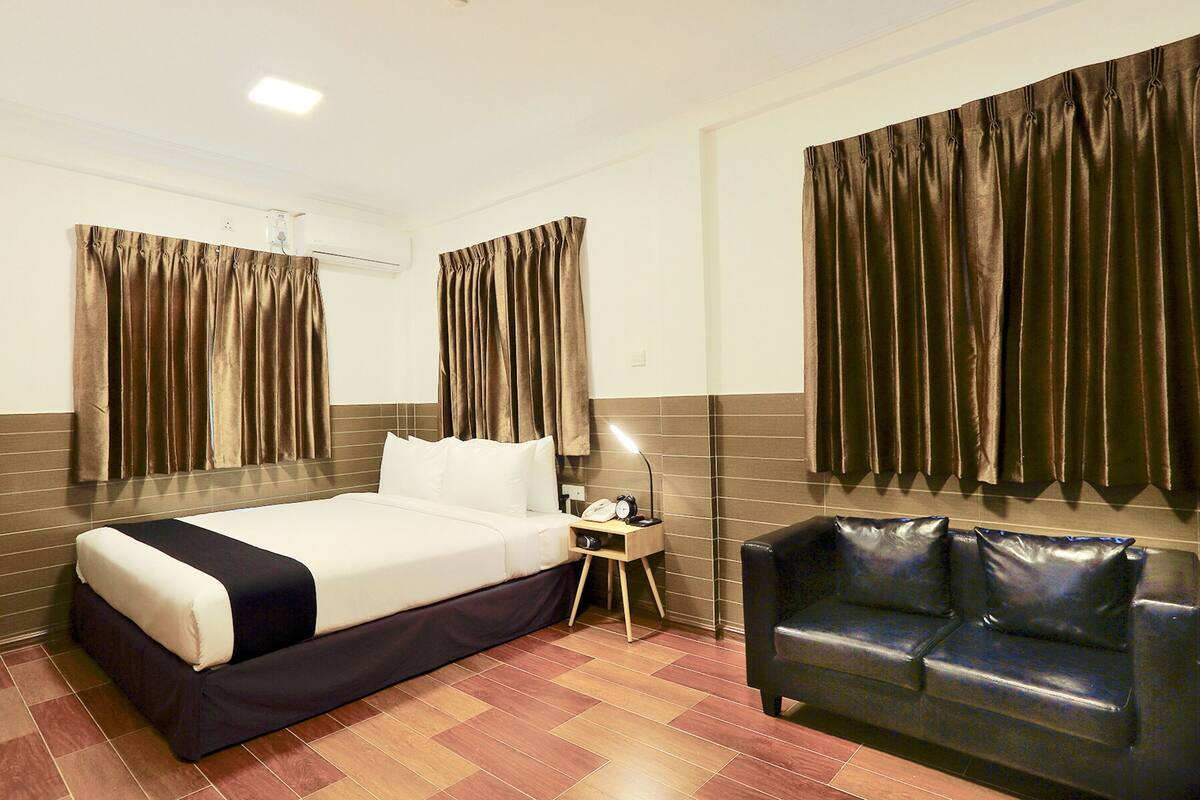 Quarto de hotel com cama, mesa de cabeceira, abajur e um sofá de couro preto.