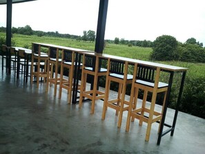 A covered outdoor seating area with wooden tables and chairs, surrounded by a concrete floor and a view of a green field. - B3 Hotel (Nakhon Si Thammarat)