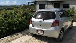 A silver car parked in front of a house with a metal roof, surrounded by greenery and a clear sky. - Villa Sur Colline Antigua (St. Johns)