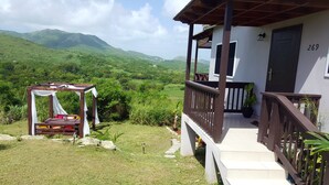A wooden gazebo with a bed and a table, a balcony with a railing, and a view of green hills. - Villa Sur Colline Antigua (St. Johns)