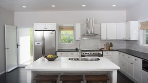 A modern kitchen with white cabinetry, a stainless steel refrigerator, a gas stove, and a large island with stools. - Villa Sur Colline Antigua (St. Johns)