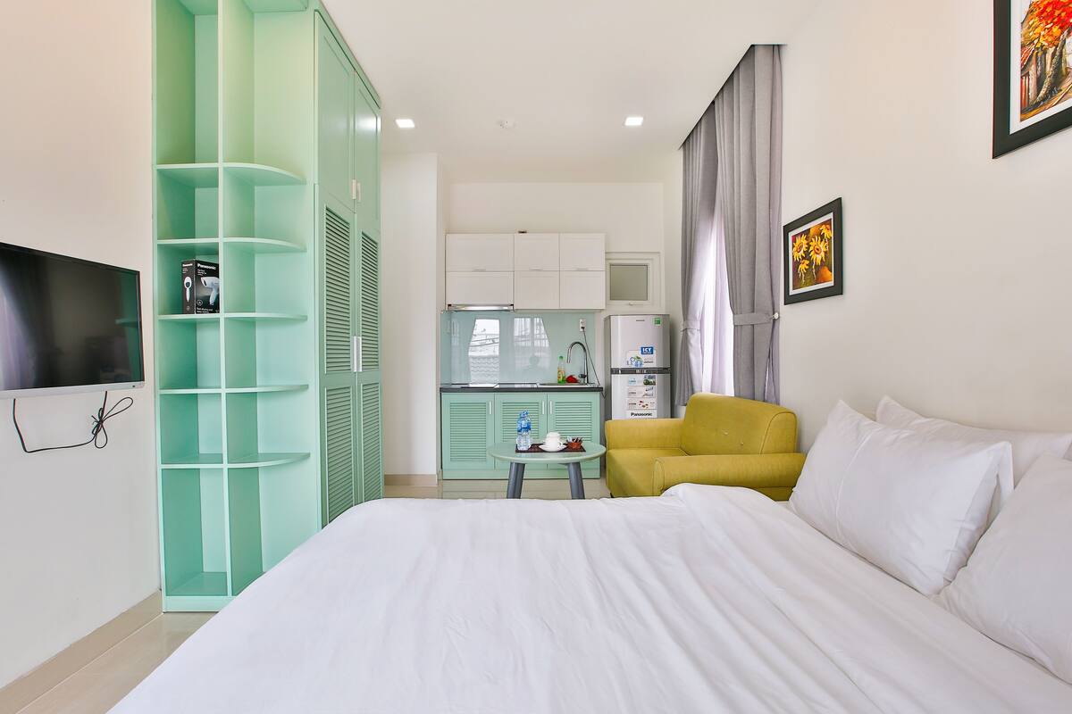 Habitación de hotel con una cama, un sillón amarillo, una mesita con una botella de agua, un televisor, un armario verde y una kitchenette al fondo.