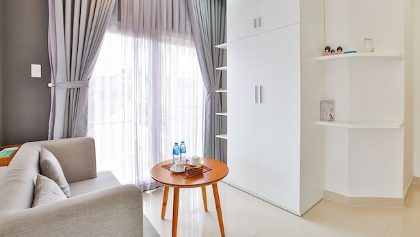 A modern hotel room with a sofa, a round wooden table with a tray of water bottles, a white wardrobe, and a shelf with decorative items. - Sunflower Boutique Studio Apartment (Ho Chi Minh City)