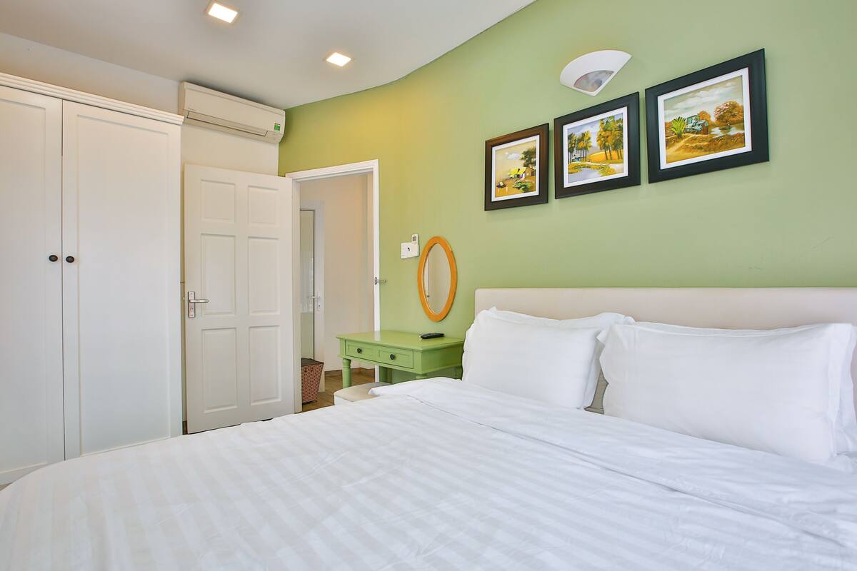 Un dormitorio con cama, una mesita de noche verde, un espejo y tres cuadros en la pared.