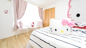 A bedroom with a large Hello Kitty bedspread, a window with curtains, a bedside table with a Hello Kitty lamp, and a wardrobe.