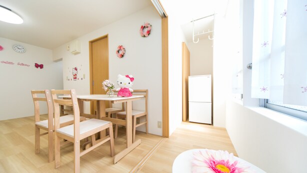 A bright room with a wooden table and chairs, a small white table with a Hello Kitty decoration, and a window with a view of the outside.